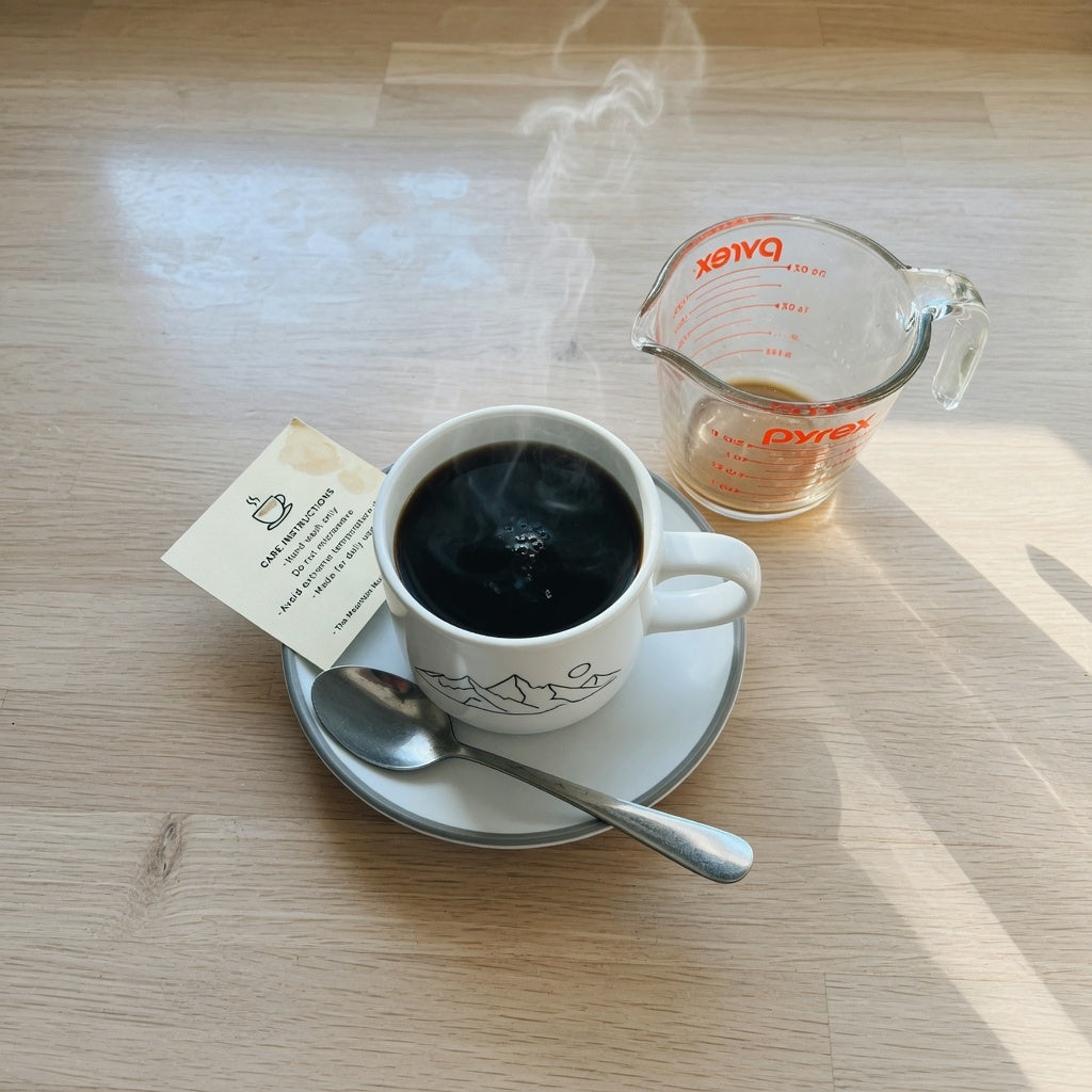Great Mountain coffee mug checklist in use: ceramic mountain-design mug beside an ounce-marked measuring cup and a printed care card on a kitchen counter