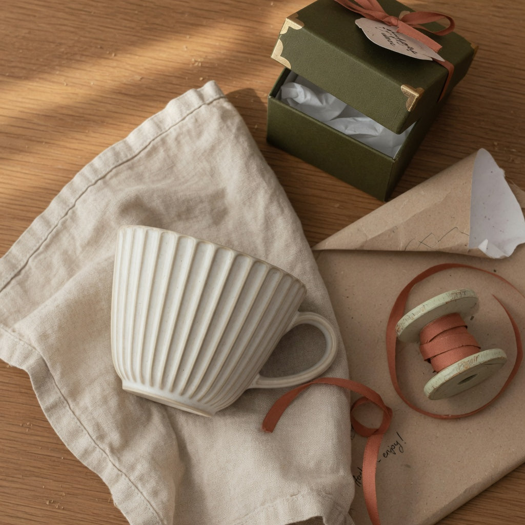Pleated coffee cup gift ready for wrapping — a fluted ceramic cup beside kraft paper, ribbon, and a small gift box