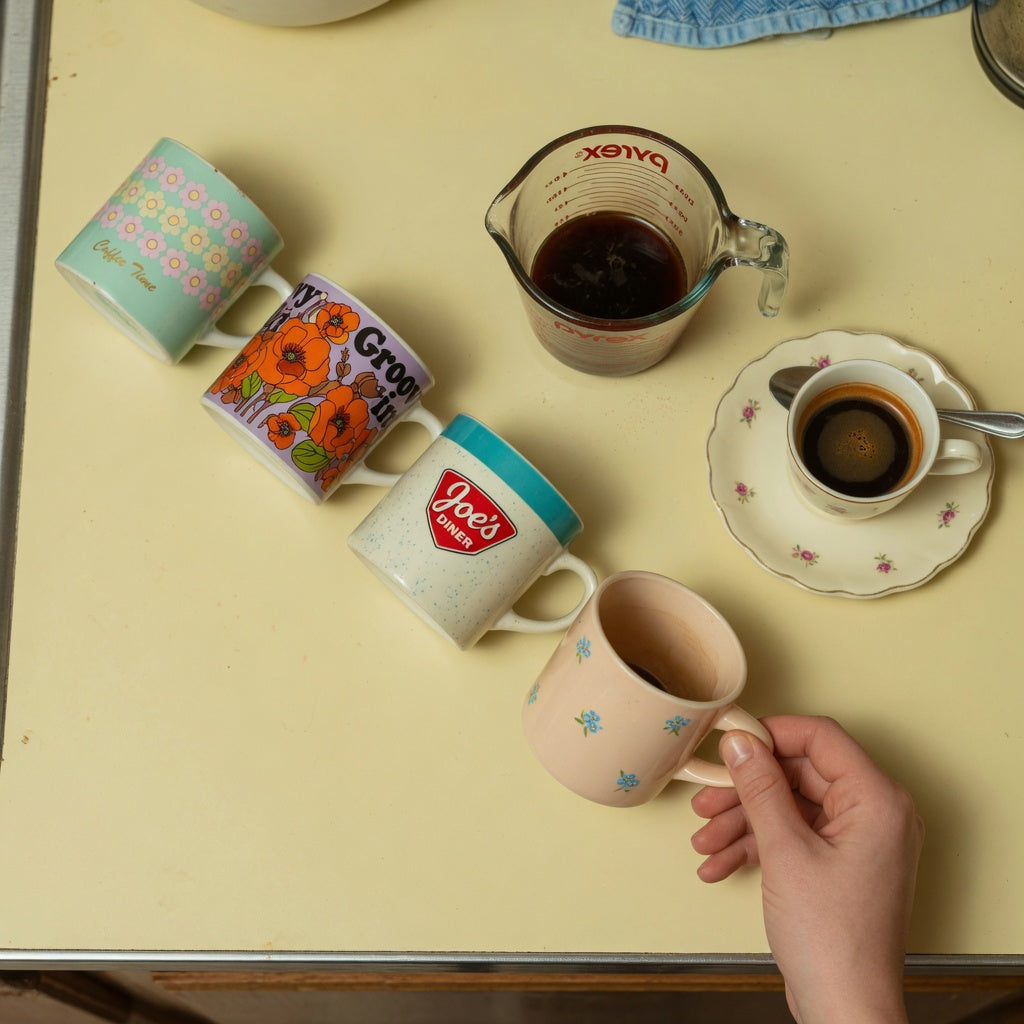 Small retro coffee mugs in pastel, floral, and diner styles beside a measuring cup and a demitasse cup on a saucer