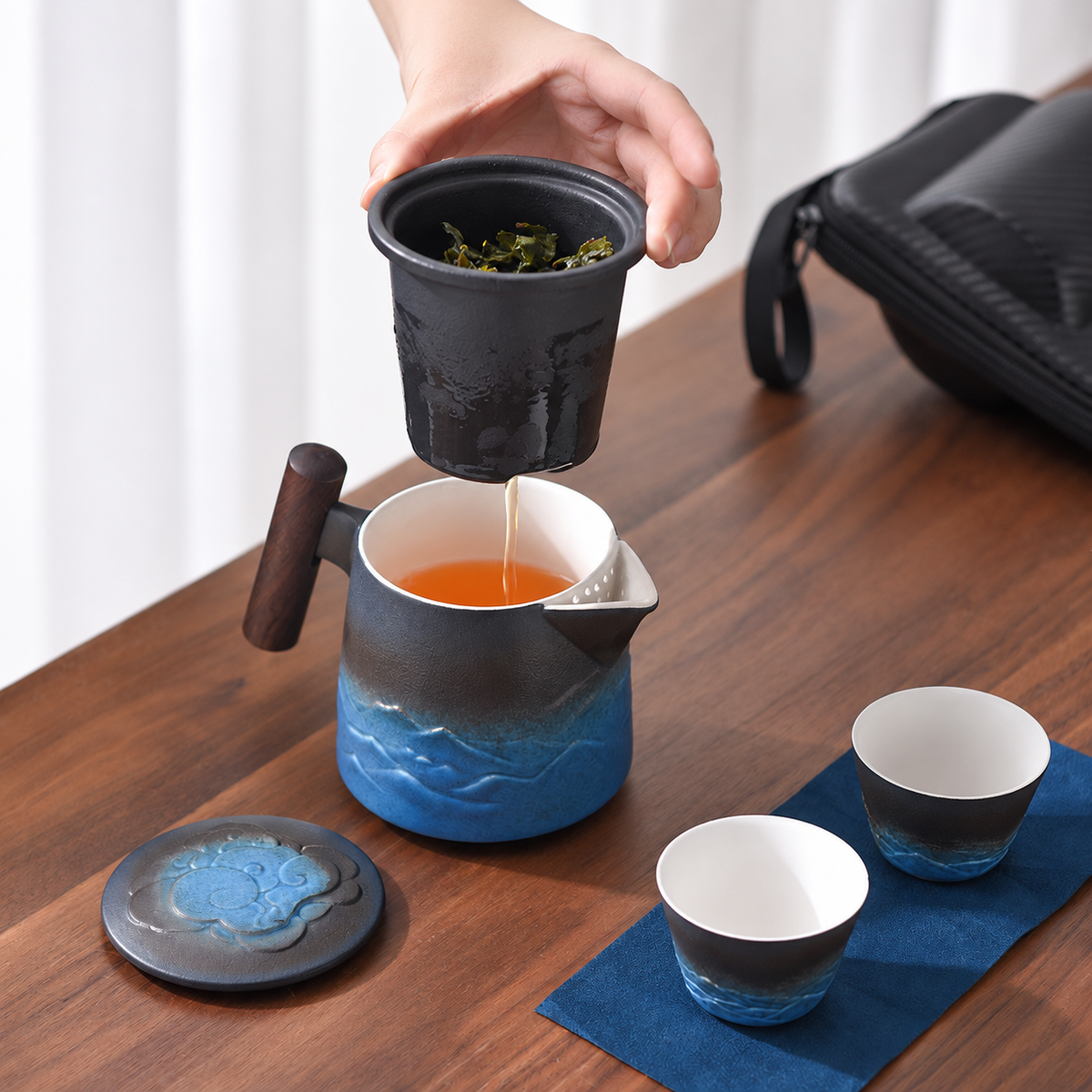A hand lifts the infuser above the black and blue travel teapot beside two matching cups
