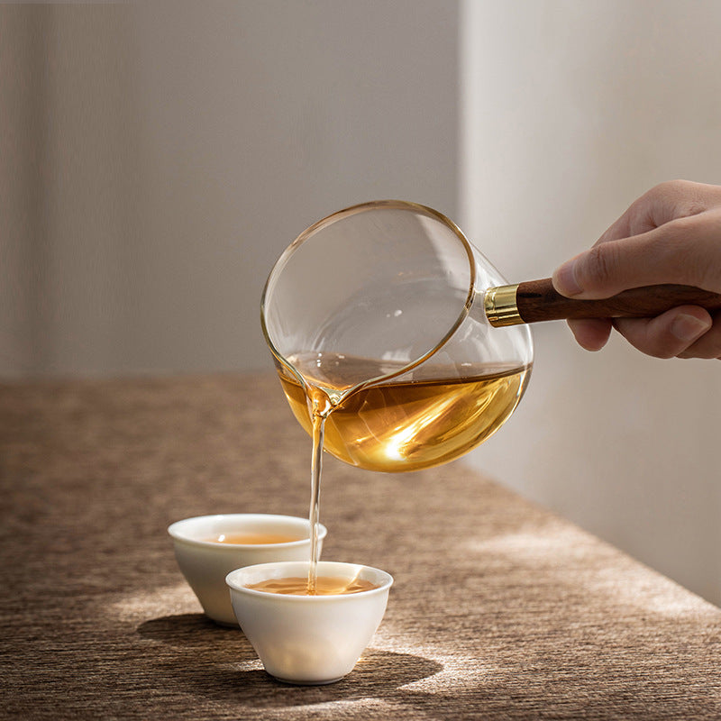 Hand pouring tea from the glass side-handle brewer into a white ceramic cup