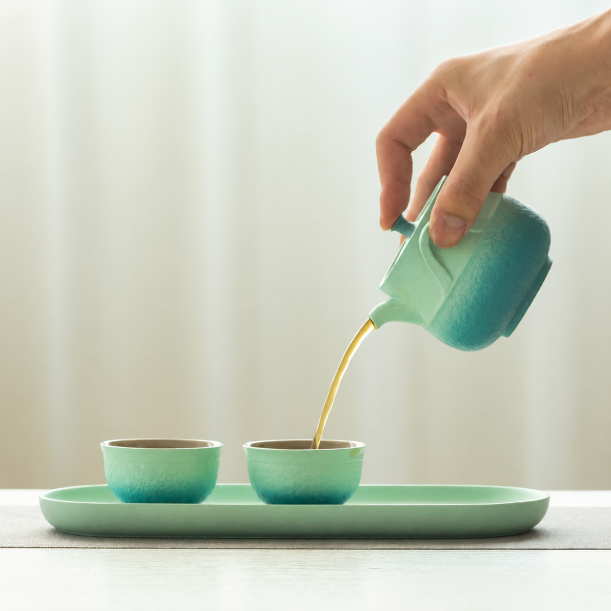 Pouring tea from a teal gradient ceramic teapot into matching cups on a tray
