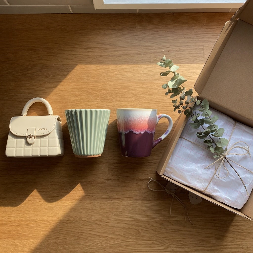 Three cute unique ceramic coffee mugs for gifts — handbag-shaped, pleated, and gradient glaze — lined up on a kitchen counter beside gift wrap
