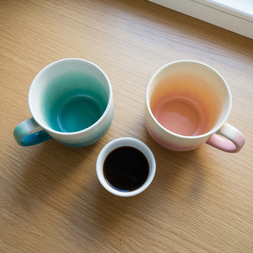 Gradient coffee mug comparison: two ceramic mugs with teal-to-white and blush-to-cream fades on a sunlit kitchen counter