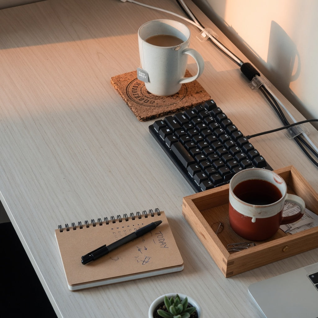 Home office coffee mug desk setup with a tall mug on a cork coaster and a round mug in a corner tray beside a keyboard
