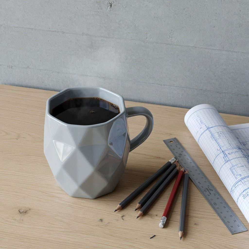 Faceted rhombus mug gift for coffee lovers styled on a minimalist architect's desk beside drafting pencils and a rolled blueprint