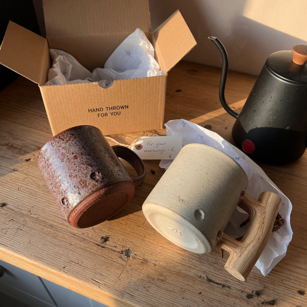 Two speckled rustic ceramic mugs for gifts on a wooden kitchen counter beside a kraft gift box and coffee kettle