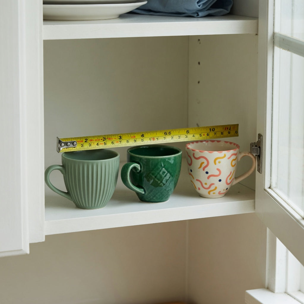 Small ceramic coffee cup storage fit guide showing Pleated, Emerald, and Retro cup shapes on a measured cabinet shelf