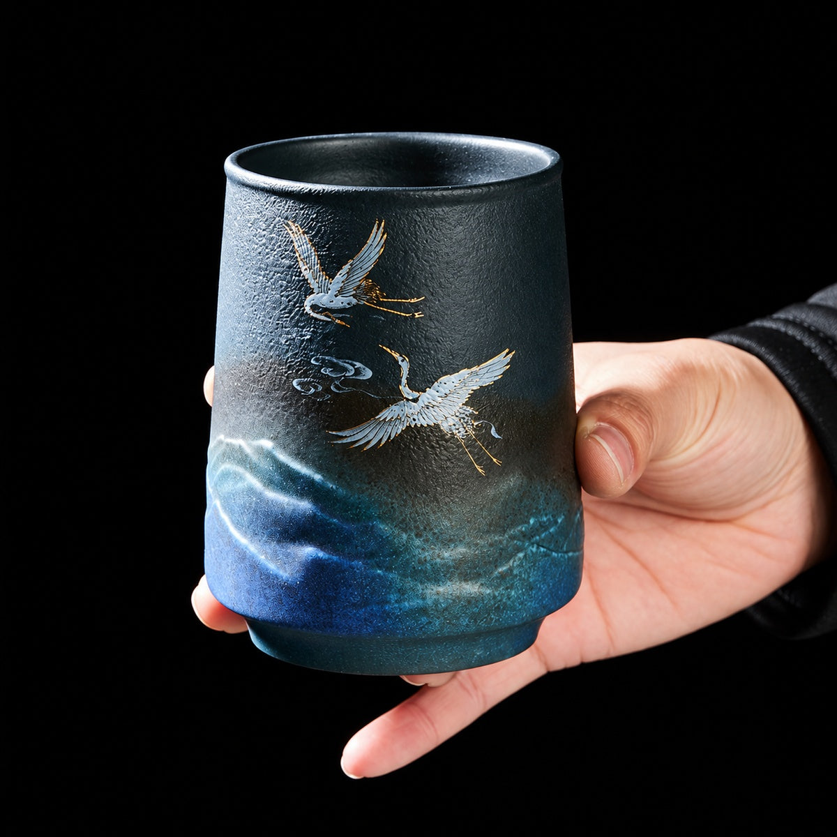 Deep blue stoneware mug held in hand showing textured glaze and crane artwork