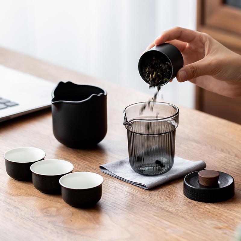 Hand adding loose leaf tea to the detached glass brewer beside the black ceramic server and three cups
