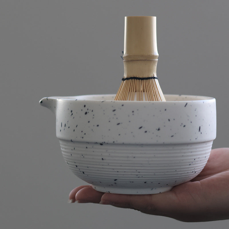 Hand supporting the speckled white matcha bowl with a bamboo whisk shown for scale and use