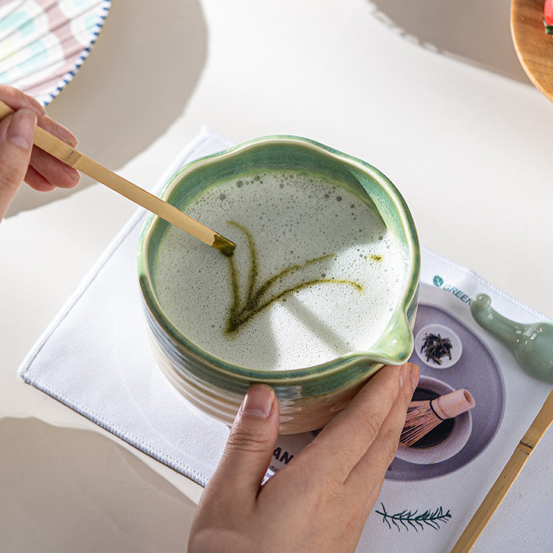 Hands decorating prepared matcha in the green scalloped-rim bowl, showing its interior and spout